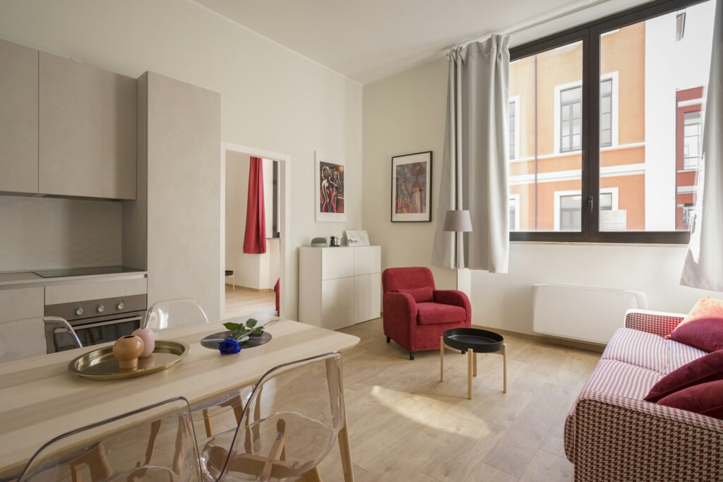 Compact studio apartment with kitchenette, small dining table, red armchair, and striped sofa by a large window