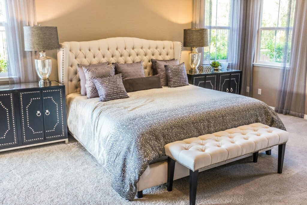 Bedroom with a tufted upholstered headboard, layered grey and cream bedding, and an upholstered bench